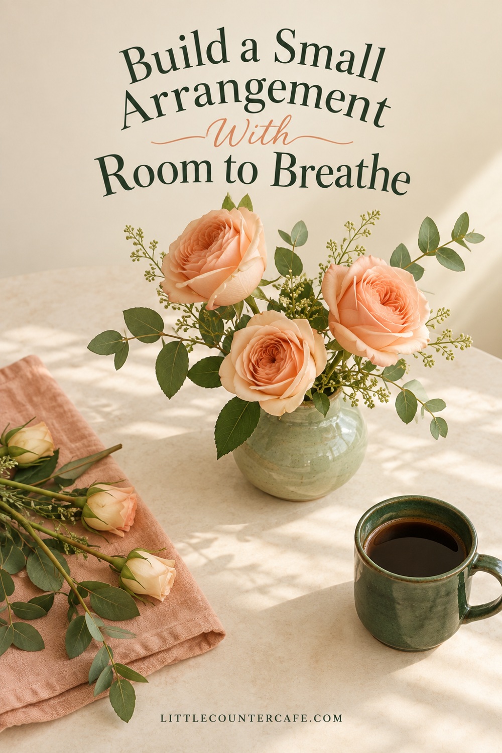 Three peach roses arranged in a sage vase on a cream tabletop beside spare stems and a green coffee cup.
